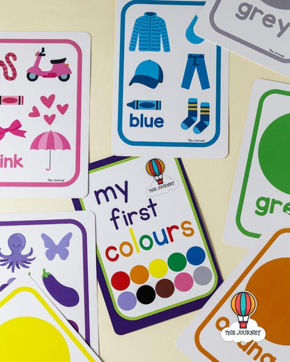 A set of colorful children's educational cards laid out on a light cream surface. The cards feature simple illustrations and words such as blue, pink, green, yellow, black, white, and grey, with one card reading 'my first colours' and another showing a scooter and hearts.
